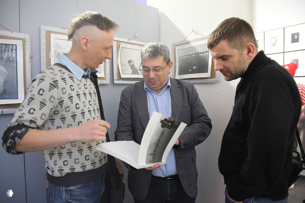 Три человека обсуждают развёрнутый фотокаталог в зале выставки; на стенах вокруг видны черно‑белые и плёночные снимки ручного оформления.
