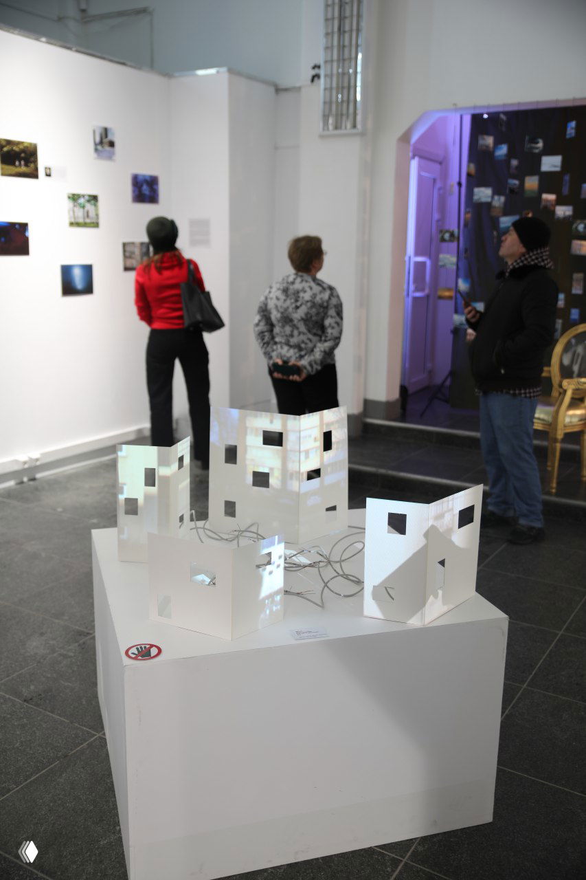 Инсталляция на белом пьедестале — бумажные макеты с проекцией, посетители осматривают работы в просторном холле арт‑пространства «РОСТ».