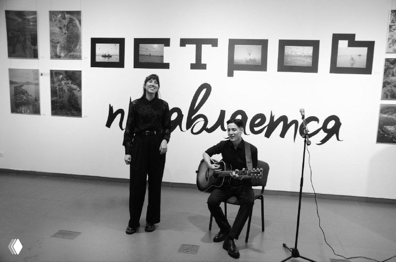 Черно‑белый кадр с музыкантами перед графическим баннером «Остров проявляется» — сценка вернисажного вечера в выставочном зале.