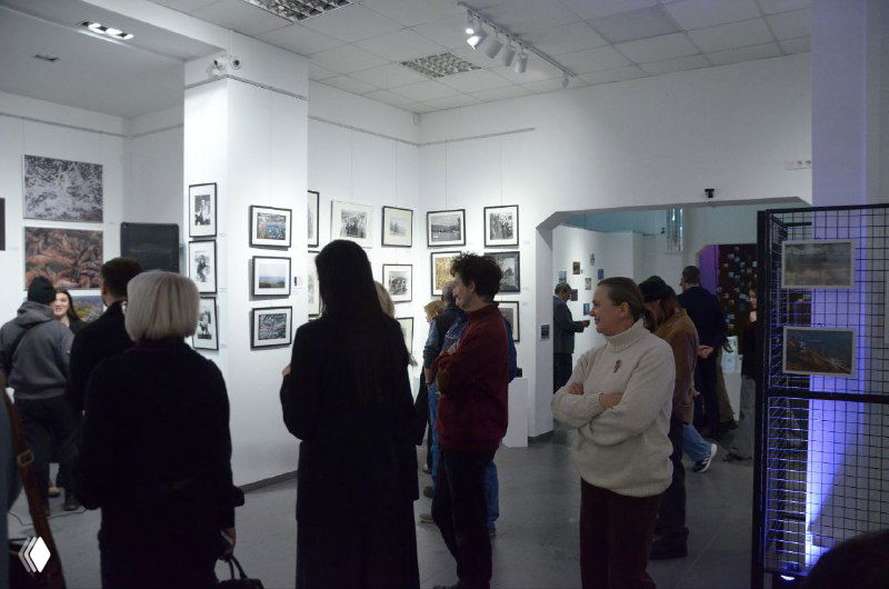 Группа посетителей осматривает фотографии на стенах галереи; кадры передают атмосферу вернисажного вечера и живого интереса публики.