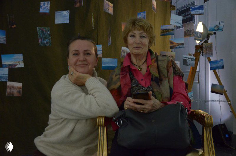 Две женщины сидят рядом у стенда с фотографиями и инсталляцией, улыбаются и наблюдают за происходящим на открытии выставки.
