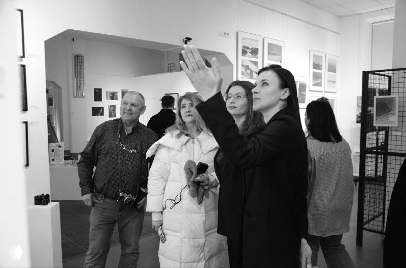 Группа людей в галерее поднимает взгляд к фотографиям, размещённым высоко на стенах; светлый зал и экспозиционный формат видны на переднем плане.