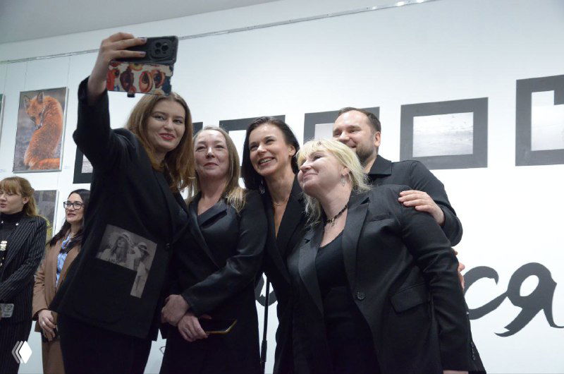 Компания гостей делает селфи на фоне стены с логотипом и кадрами выставки — живой момент открытия и неформального общения посетителей.