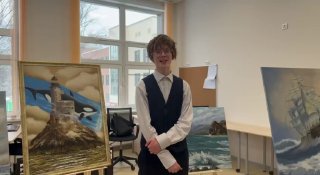 Артём Захарченко в школьном классе стоит рядом с мольбертами, на которых экспонируются масляные морские пейзажи; видны окна и интерьер аудитории.