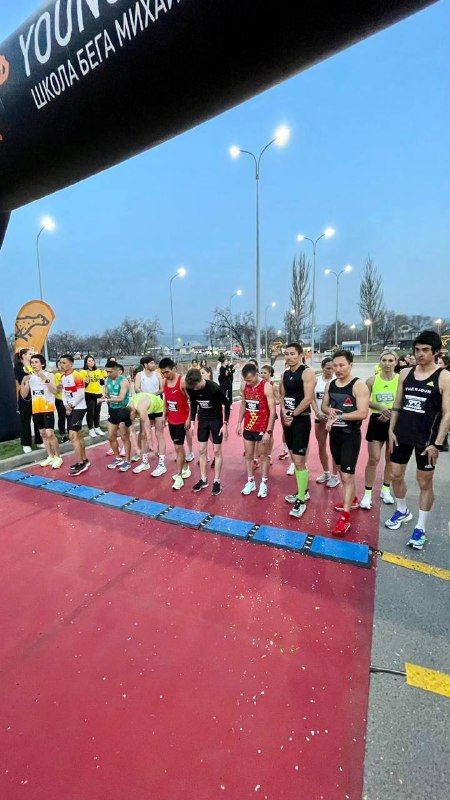 Стартовая сцена Spring Race: группа юных бегунов в стартовой области под аркой YoungRunner вечером, участники в форме и с номерами готовятся к забегу.