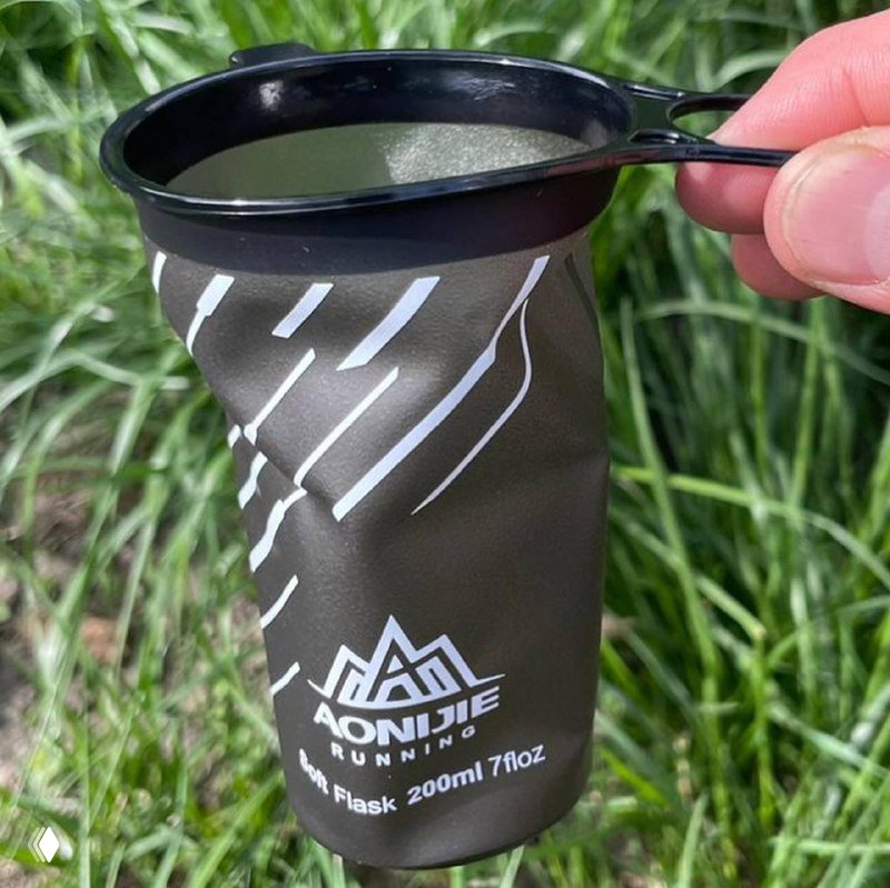 Рука держит мягкий многоразовый стаканчик Aonijie на фоне зелёной травы, видна петля-держатель и открытый край — удобство использования на трассе.