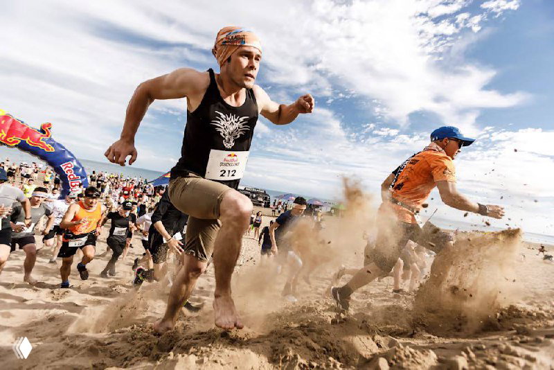 Группа бегунов на старте по песку: участники в номерах выбегают по песчаному полю, клубы пыли и интенсивный старт