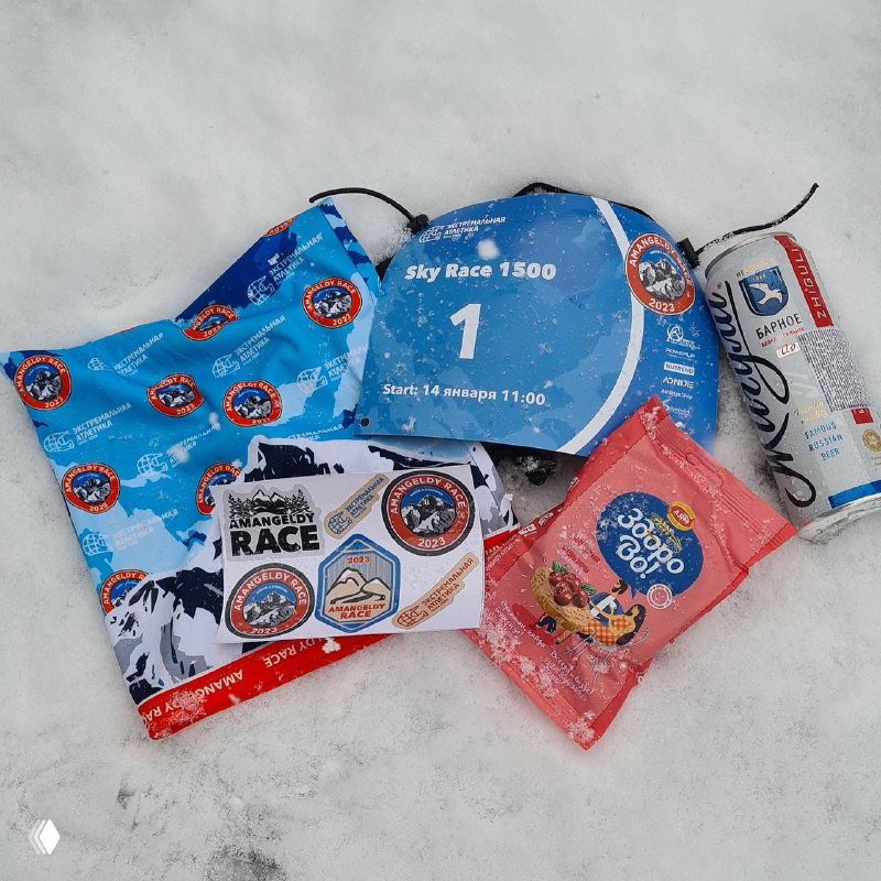 Стартовый набор AMANGELDY RACE, разложенный на снегу: нагрудный номер, брендированный баф, стикеры, хлебцы, батончик и безалкогольный напиток от партнёров.