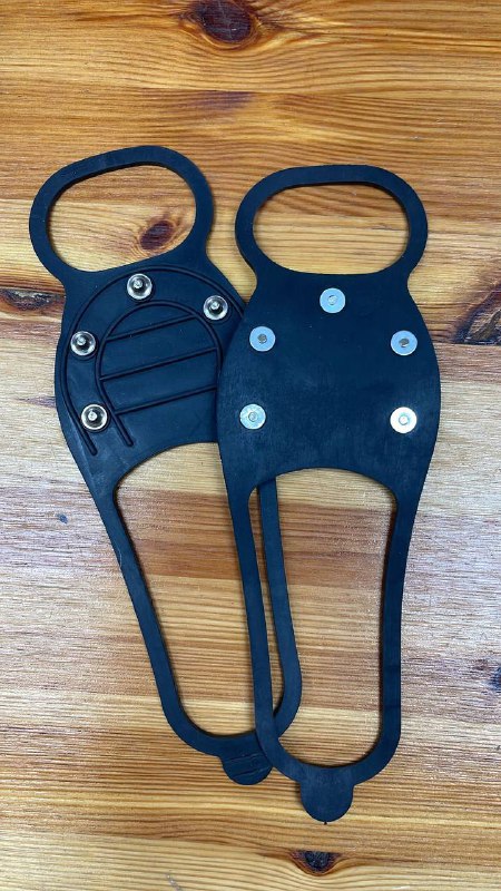 Пара чёрных ледоступов (антилёд) на деревянной поверхности, видны ремешки, подошва и металлические шипы, пример экипировки для бега по льду.