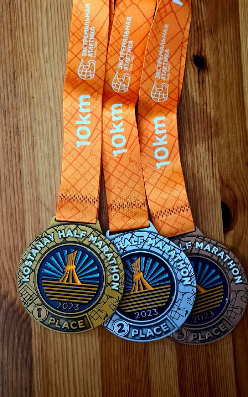 Три медали Kostanay Half Marathon на деревянной поверхности: золото, серебро и бронза с разноцветными лентами и годом 2023, общий вид комплекта наград.