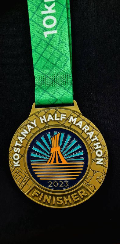 Золотая медаль-финишер Kostanay Half Marathon 2023 на ярко‑зелёной ленте; видна надпись FINISHER и стилизованный логотип забега на лицевой стороне.