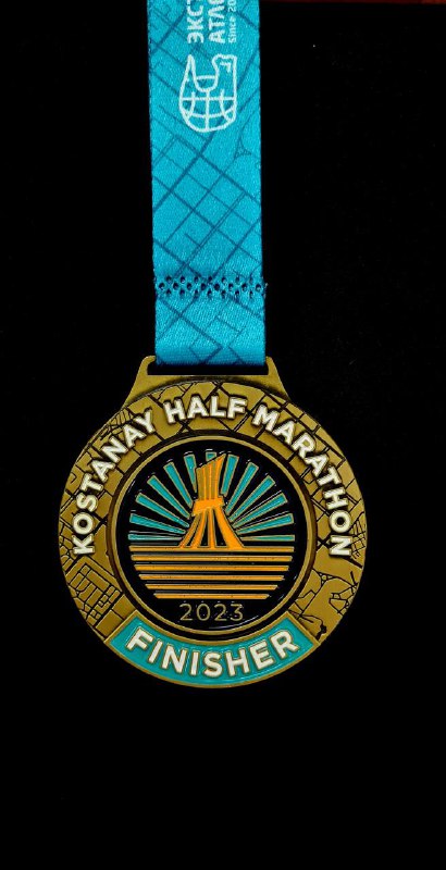 Медаль-финишер Kostanay Half Marathon 2023 на бирюзовой ленте; видно текст мероприятия и год, фон тёмный, акцент на дизайне медали.