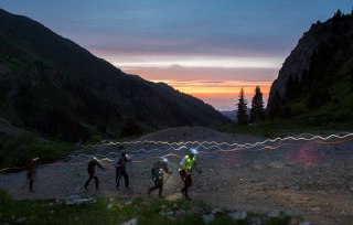 Бегуны в ночной трассе со налобными фонарями: световые следы от огней создают динамичные линии на фоне горной долины и вечернего неба