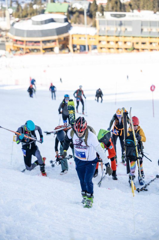 Спортсмены в плотном темпе поднимаются по снежному склону с переносом лыж на рюкзаках, видно напряжение и технику восхождения на трассе.