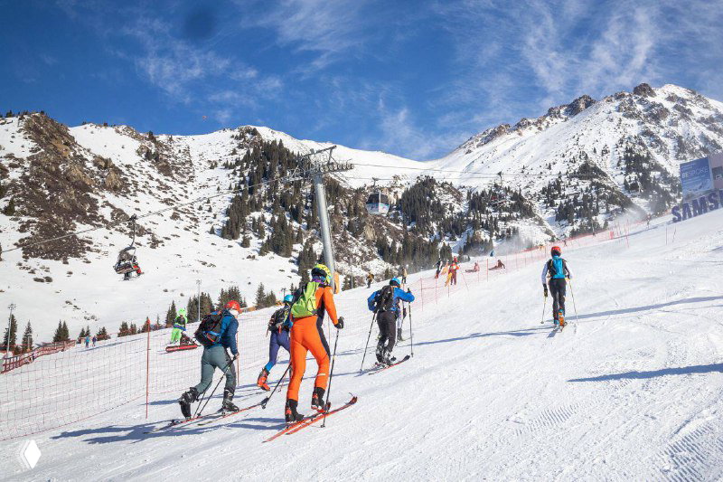 Несколько команд участников идут по ровной части трассы возле горнолыжной инфраструктуры курорта, видны маркировка и защитные сетки.