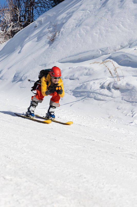 Младший участник в красном шлеме на компактных лыжах выполняет техничный спуск по снежному склону с выраженной концентрацией.