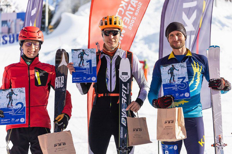 Победители и призёры на подиуме с дипломами и призами: трое спортсменов держат плакаты и кубки на фоне баннеров мероприятия.