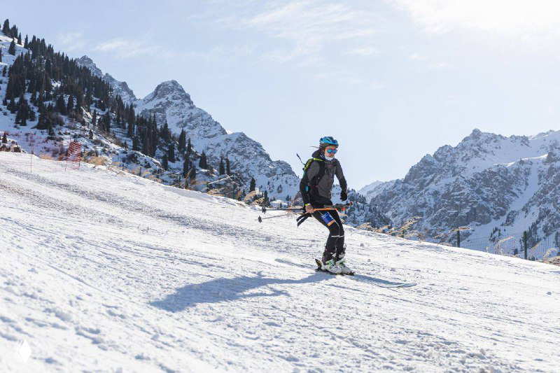 Одиночный спортсмен на фоне заснеженных вершин идет по пологому склону в горной местности, видны трек и яркое дневное освещение.