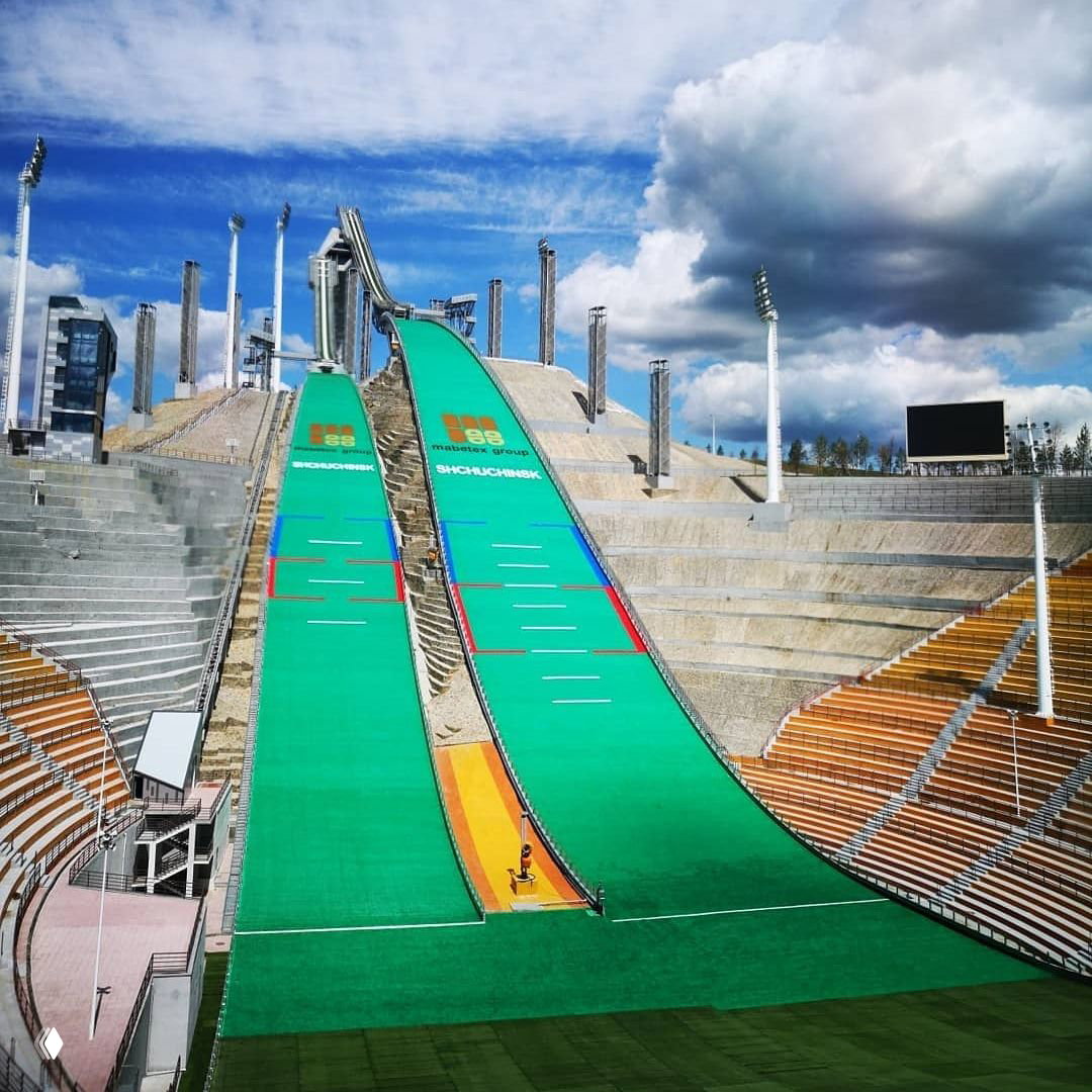 Фото трамплина в Щучинске: две широкие зелёные наклонные трассы для прыжков и старта Red Bull 400, трибуны и облачное небо на заднем плане.