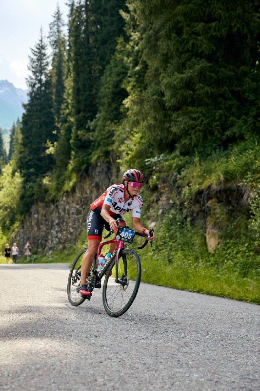 Шоссейная гонщица в красно-белой экипировке ускоряется по горной дороге, позади видны скалистые обрывы и лесистый склон.
