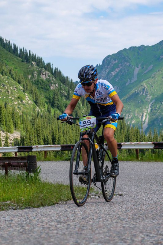 Участник на горном велосипеде с номером 199 проезжает по трассе в горах, на заднем плане зелёные долины и скалистые пики.