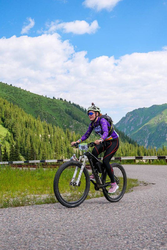 Женщина на MTB в фиолетовой куртке едет по ровной дороге в горах, вокруг луг и зелёная долина под частично облачным небом.