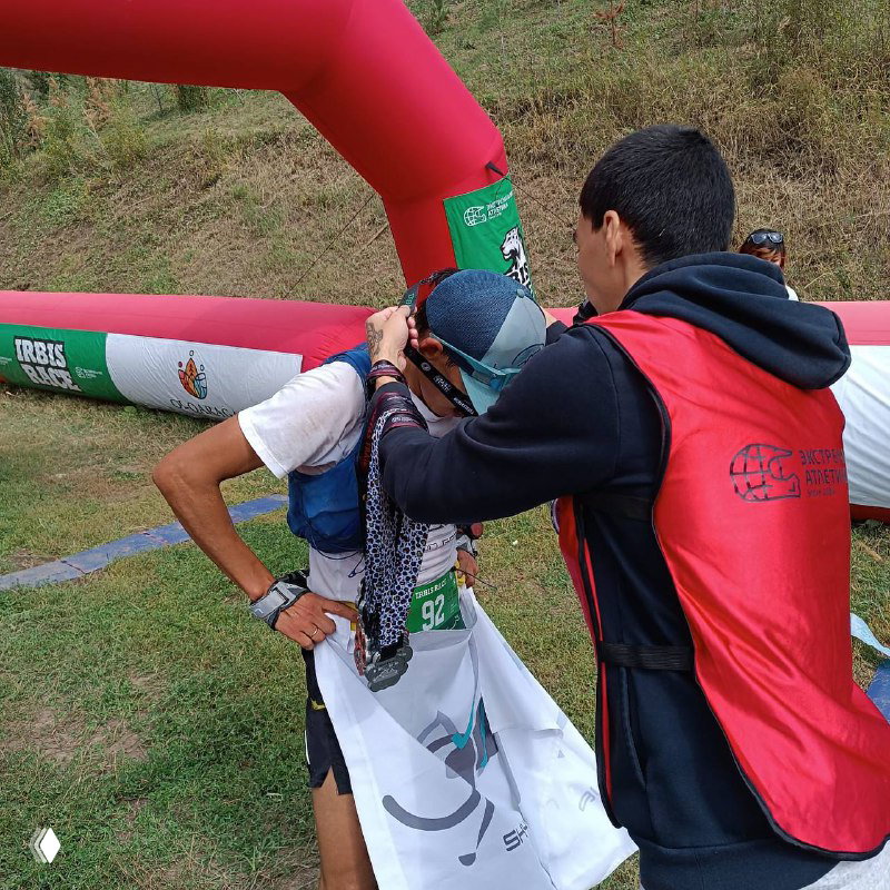 Волонтёр надевает медаль участнику у финиша Irbis Race; видна надувная арка и зрители на фоне горной трассы.