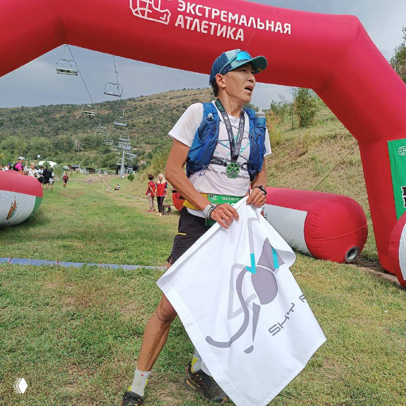 Участник с флагом и медалью стоит у финиша Irbis Race, позади видна надувная арка и холмистый ландшафт соревнований.