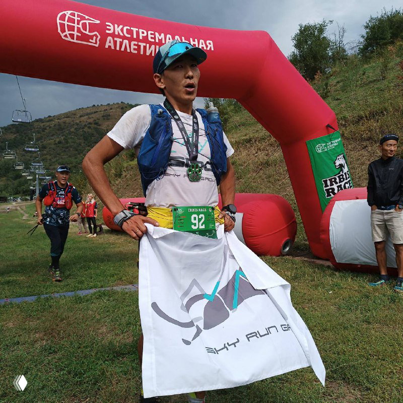 Финиширующий спортсмен держит флаг и медаль у арки «Экстремальная Атлетика» на травянистом склоне, кадр близкого плана.