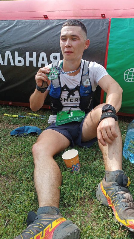 Сидящий на траве финишер отдыхает с медалью и бумажным стаканчиком, грязная экипировка и номер на поясе отчётливо видны.