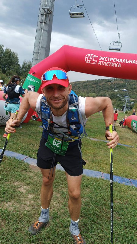 Улыбающийся финишер с трековыми палками и номером 62 позирует у финишной арки на горной трассе забега.