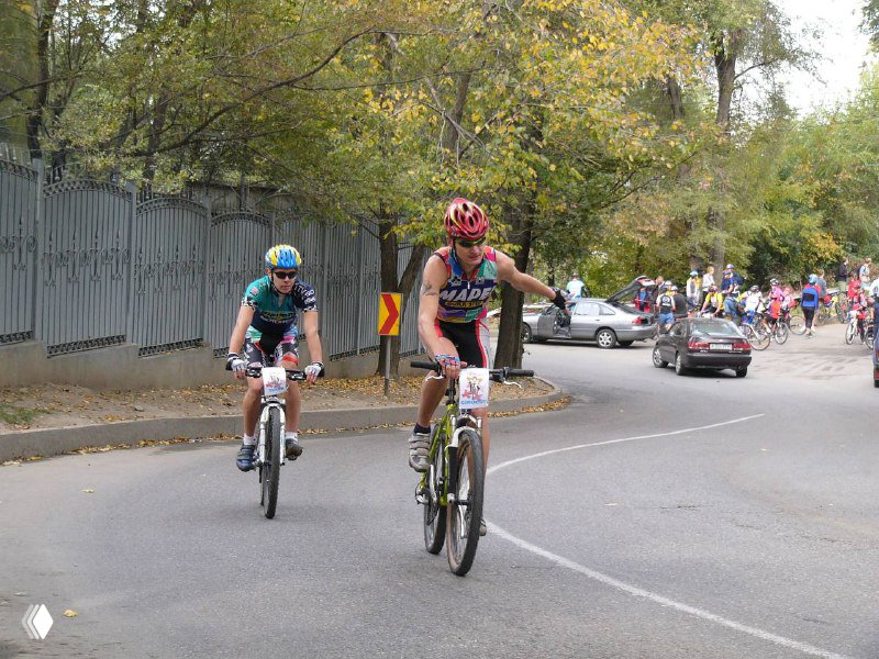 Два лидера гонки движутся по городскому участку трассы: велосипеды МТБ, скорость и автомобиль на обочине.