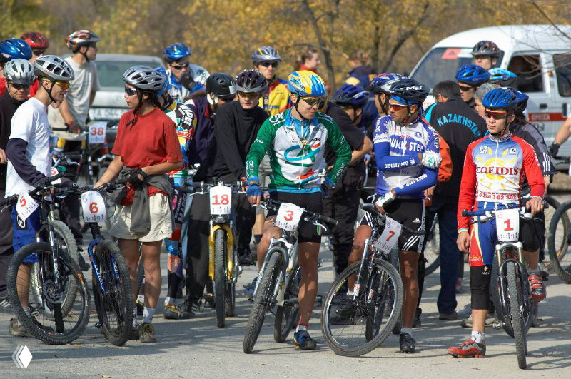 Стартовая линия гонщиков в защитных шлемах и экипировке; участники готовятся к трассе, слушают инструкции и настраиваются.