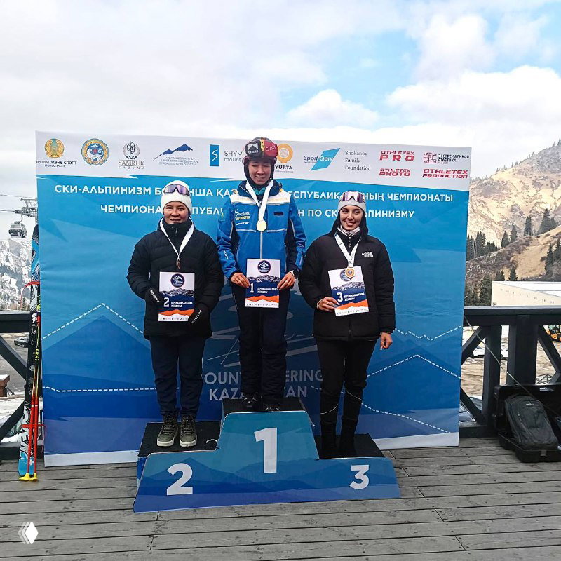 Фото женщин на пьедестале Чемпионата РК по ски-альпинизму: три спортсменки с медалями и дипломами на фоне горного баннера и неба.