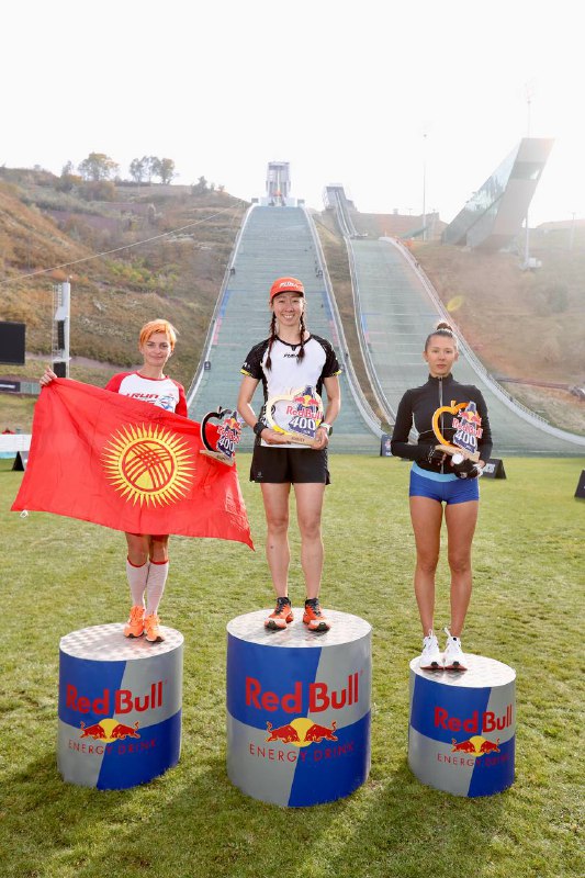 Три призёрки женского забега Red Bull 400 на подиуме; одна участница держит национальный флаг, позади виден трамплин и поле старта соревнования.