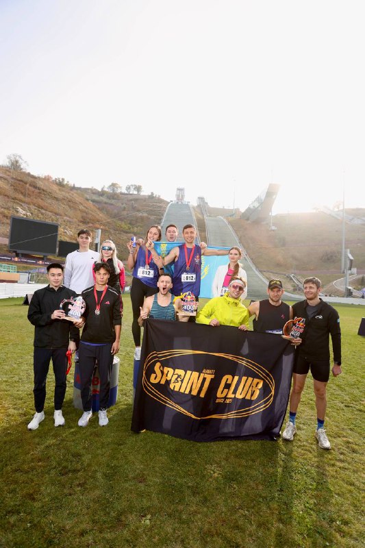 Командная фотография призёров эстафеты Red Bull 400: участники с кубками и баннером Sprint Club на травяной площадке перед трамплином.