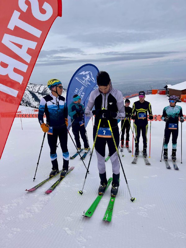 Участники на старте дисциплины Вертикальный километр рядом со стартовыми флагами и организаторами на снежной площадке курорта.