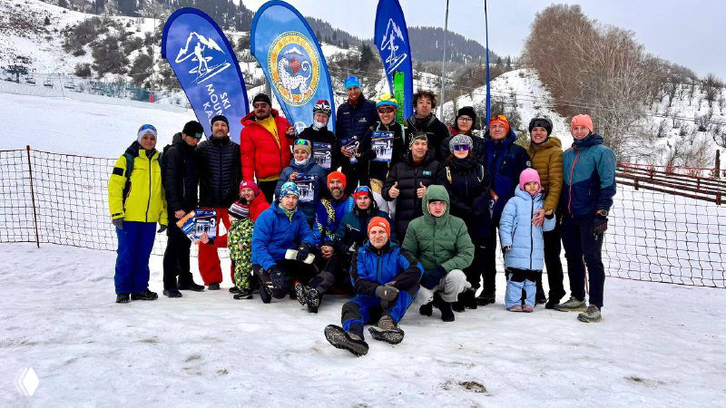 Групповое фото участников и организаторов соревнований по ски‑альпинизму на заснеженной площадке; видны флаги и зимний фон гор.