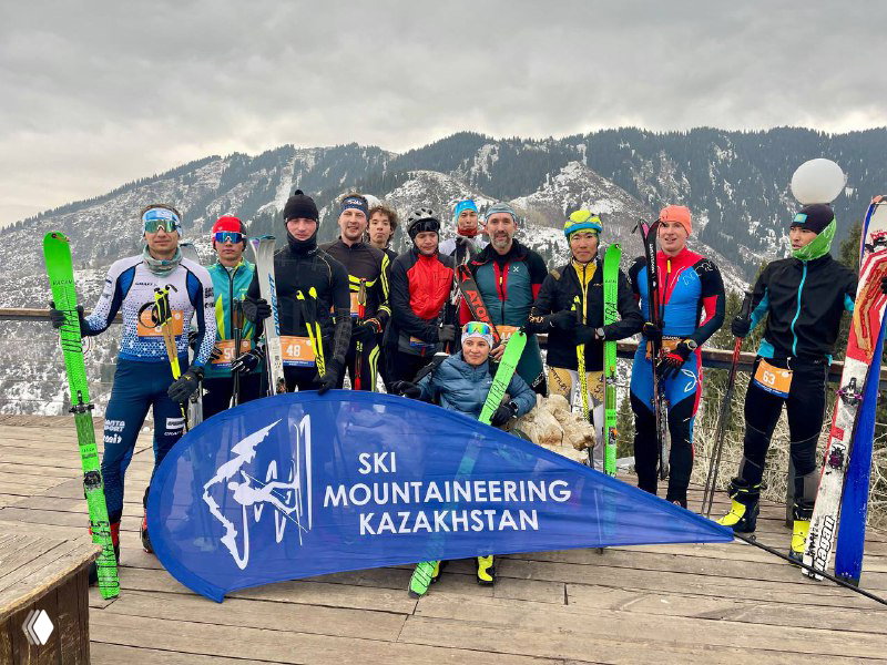 Команда лыжников с экипировкой стоит за баннером «Ski Mountaineering Kazakhstan» на деревянной платформе на фоне горной панорамы.