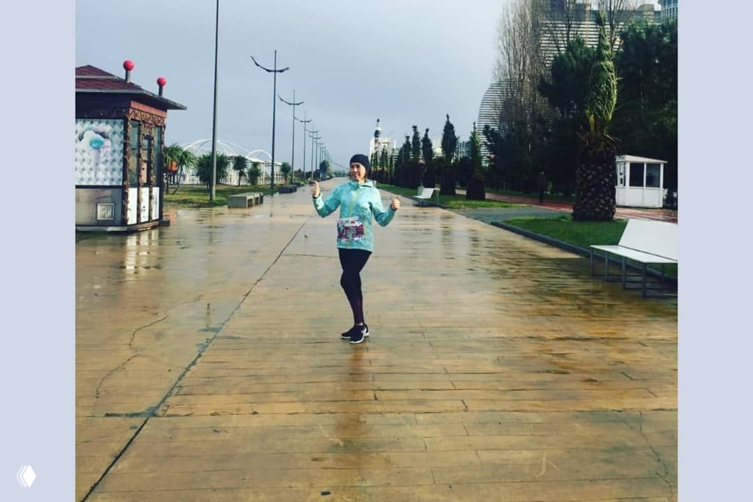 Одиночная бегунья на набережной в дождливую погоду, в спортивной куртке и с номером на талии, просторная деревянная дорожка и городской фон.