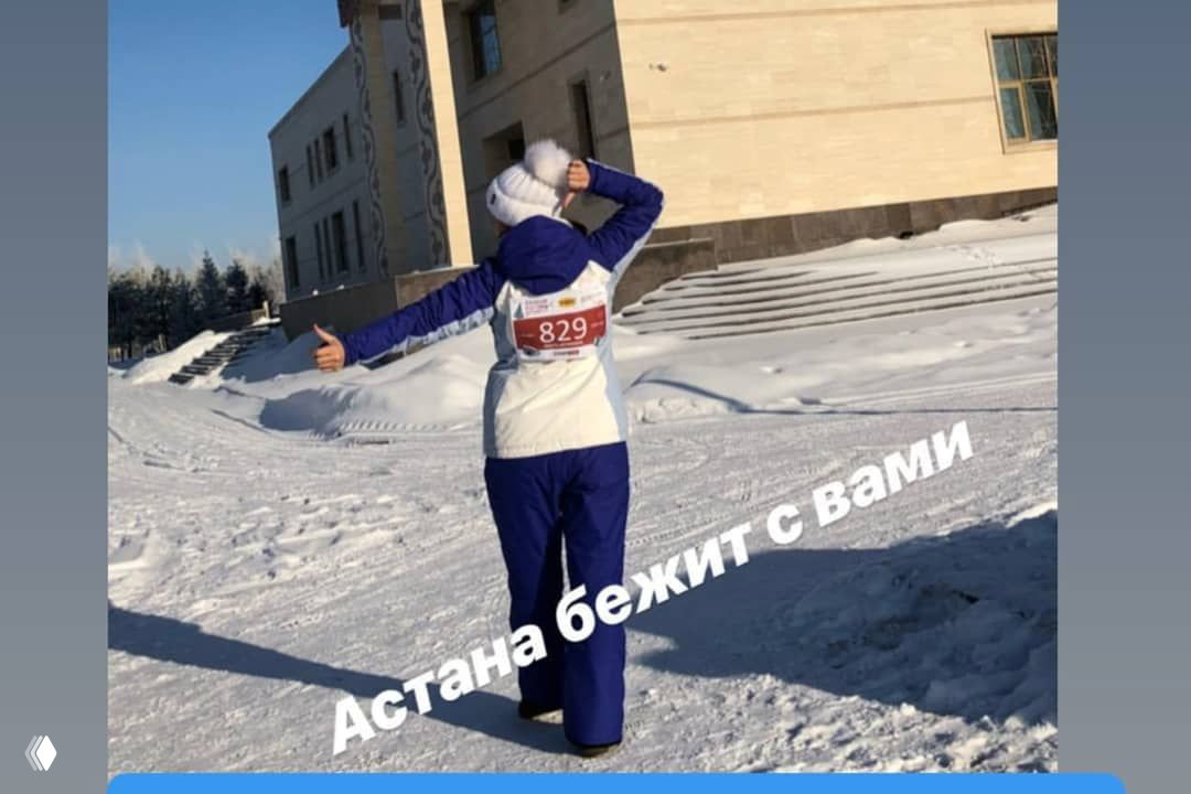 Бегунья спиной в зимней экипировке со стартовым номером 829, вокруг снежный пейзаж и здание — подпись на фото: «Астана бежит с вами».