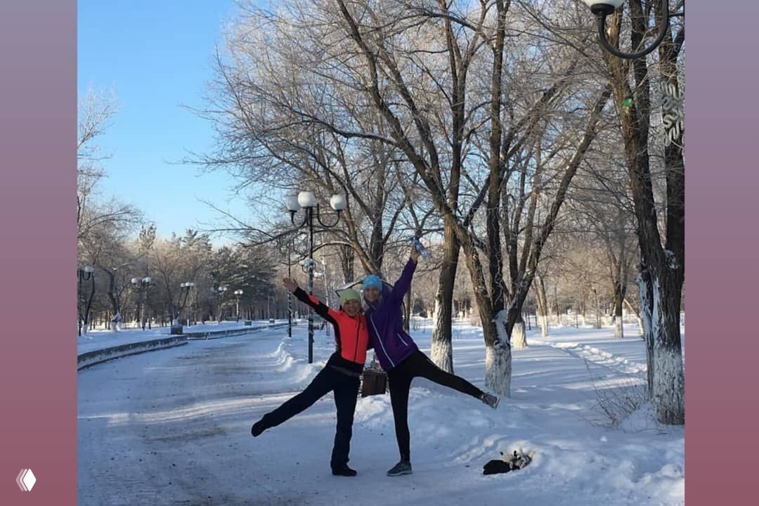 Две женщины-бегуньи в парке зимой, подняв руки и позирующие на заснеженной аллее; вокруг деревья и фонари, светлое небо.