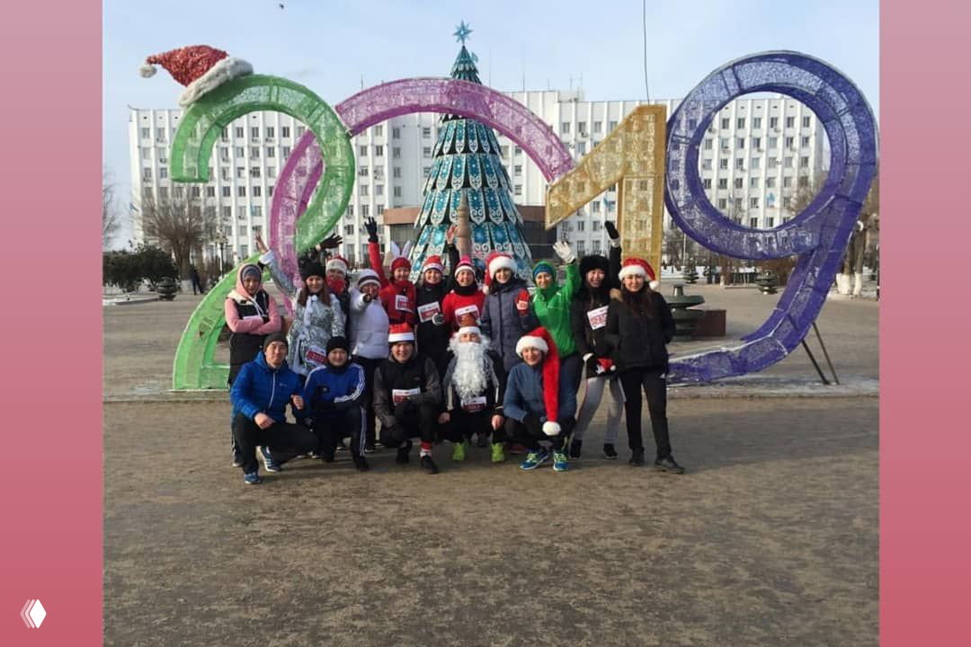 Большая группа бегунов в новогодних шапках у декоративной инсталляции «2019» и ёлки в центре города, участники позируют перед праздником.