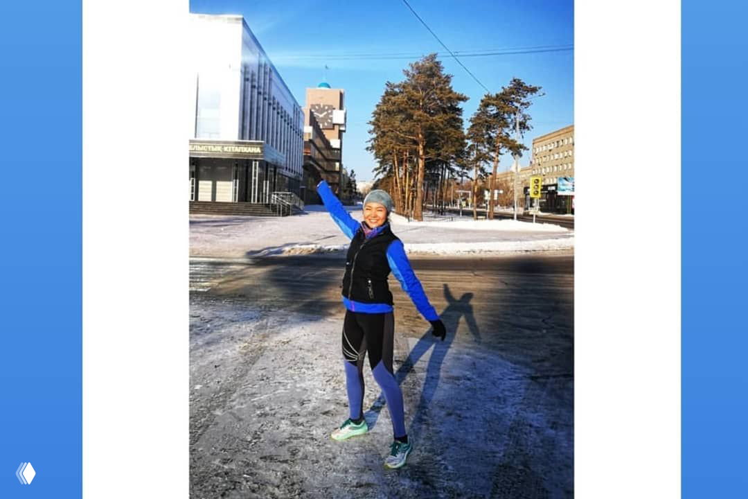 Одиночная бегунья на городской улице на фоне сосен и зданий, солнечный свет, зимняя беговая экипировка и тёплые колготки.