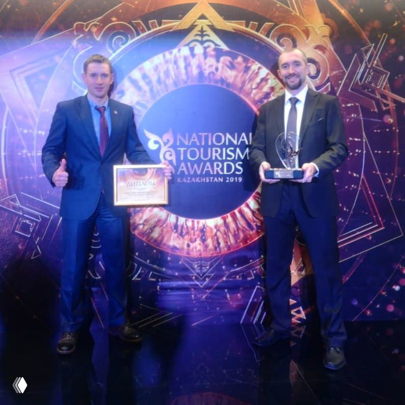 Двое мужчин в деловых костюмах держат награду и диплом на фоне яркого оформления с логотипом National Tourism Awards Kazakhstan