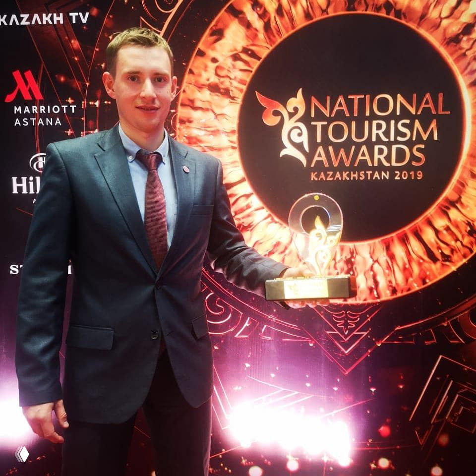 Мужчина в костюме держит статуэтку на фоне большого круглого логотипа National Tourism Awards Kazakhstan и световой декорации сцены