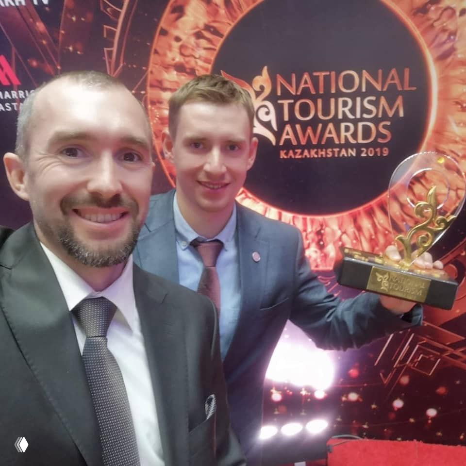 Селфи двух организаторов в смокингах на фоне логотипа National Tourism Awards Kazakhstan, видна статуэтка и праздничная атмосфера