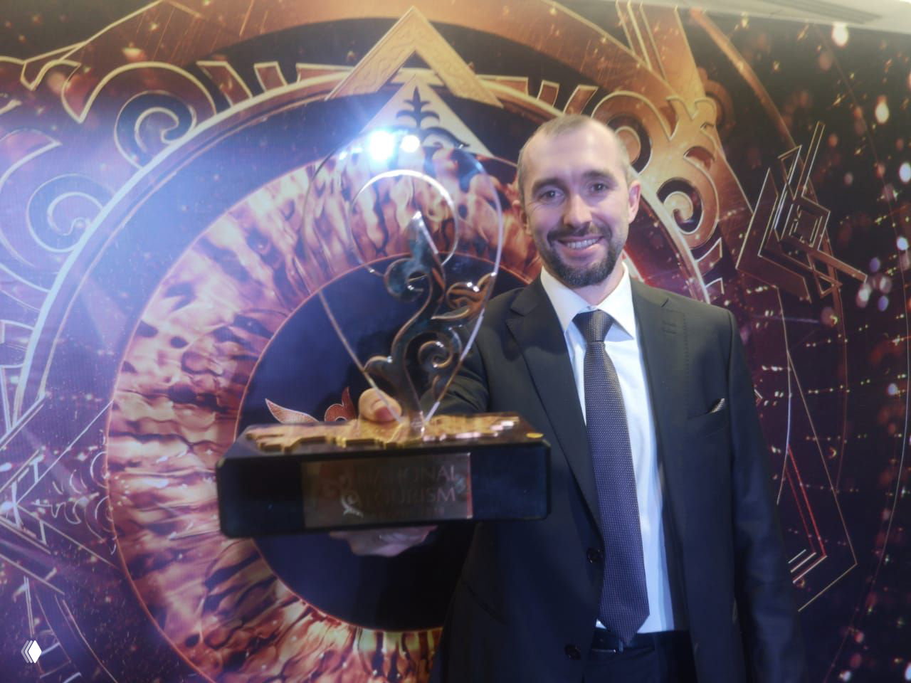 Мужчина в костюме держит статуэтку крупным планом, на заднем фоне декоративная сцена с логотипом National Tourism Awards Kazakhstan