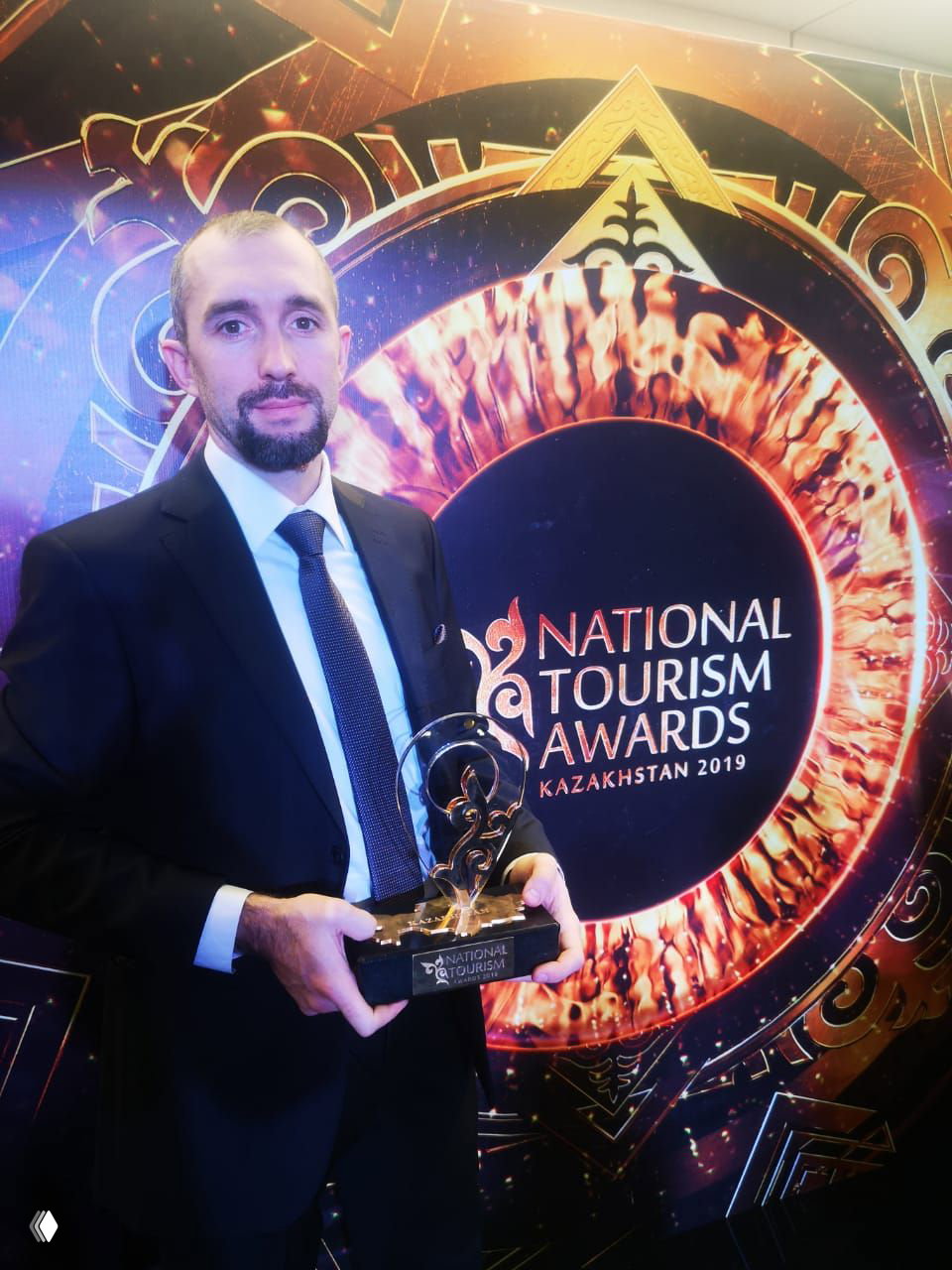 Мужчина в официальном костюме с наградой на фоне яркой оформленной фотозоны с логотипом National Tourism Awards Kazakhstan