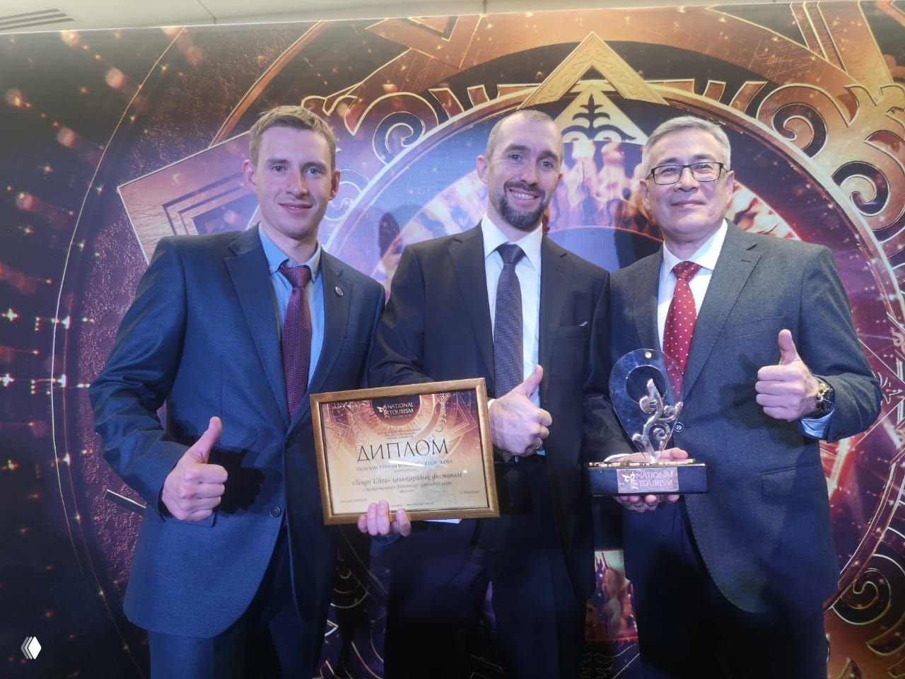 Трое мужчин в костюмах держат диплом и статуэтку на фоне оформления National Tourism Awards Kazakhstan, позируют с поднятыми большими пальцами